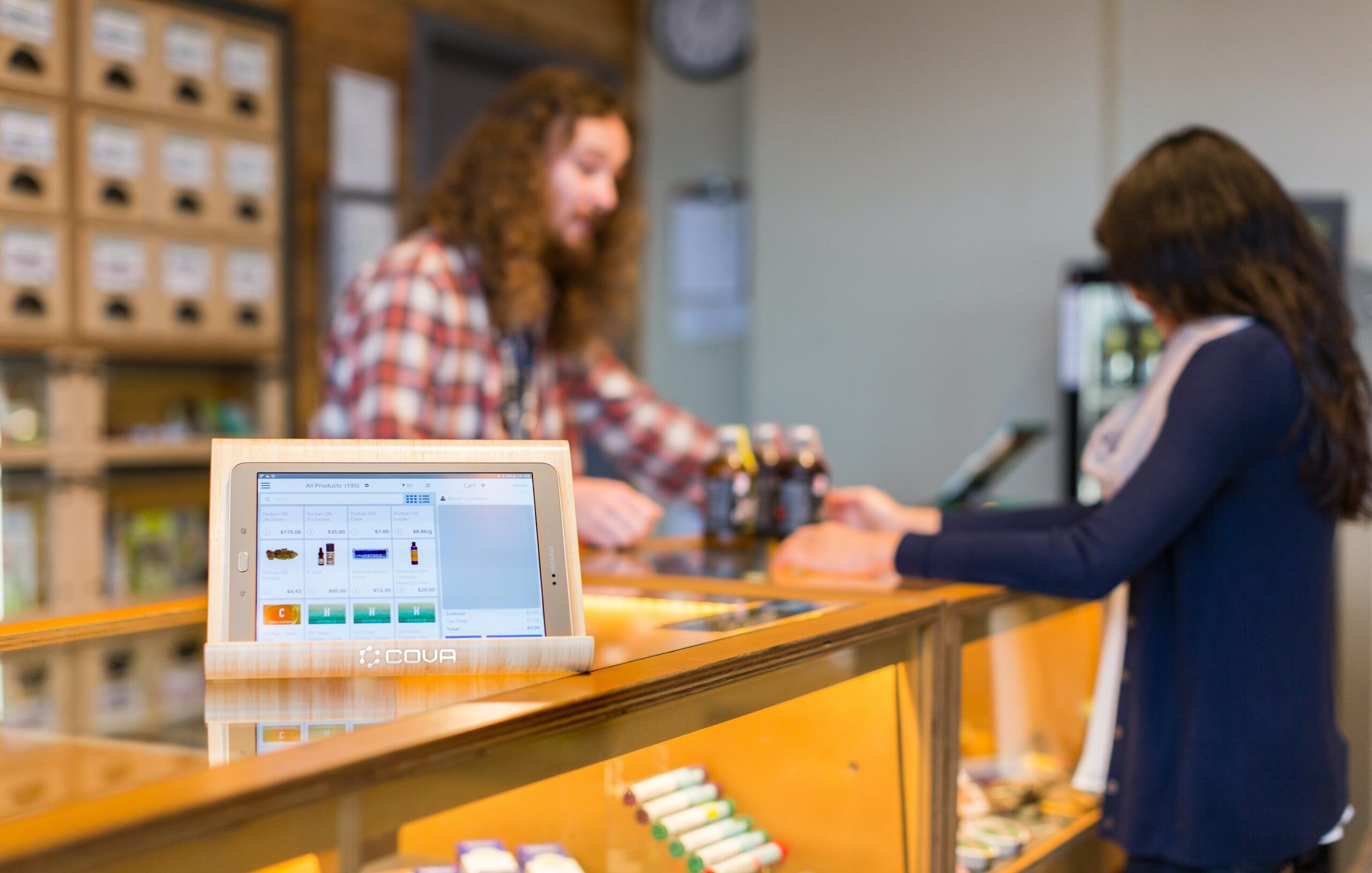 A lit-up screen displaying Cova's point of sale cannabis software is in focus atop a glass counter in a dispensary. In the background, a male budtender with long, curly hair helps a female cannabis customer choose from an array of cannabis beverages and edibles.